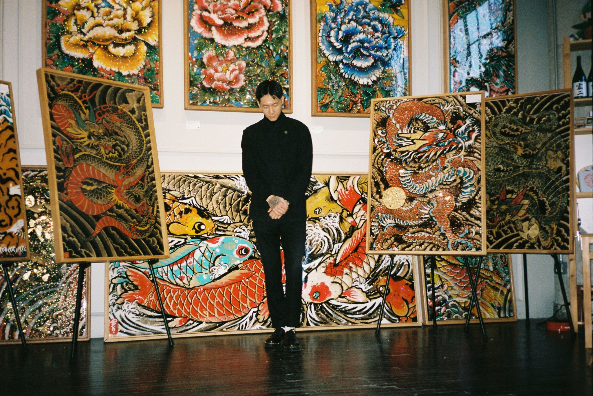Person standing in art gallery with colorful dragon and floral paintings displayed around.