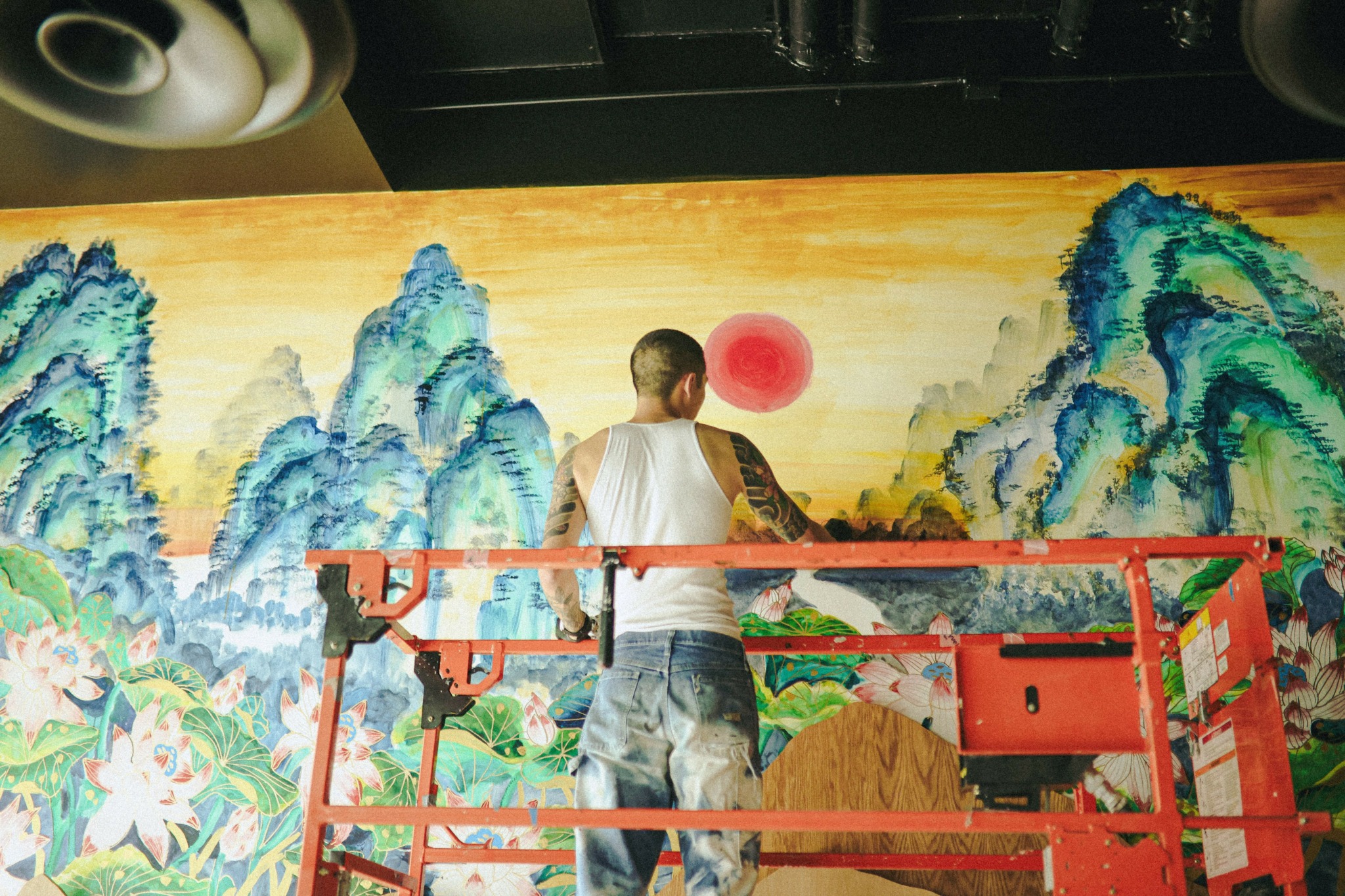 Person standing on a red scaffold in front of a colorful mountain landscape painting with a red sun.