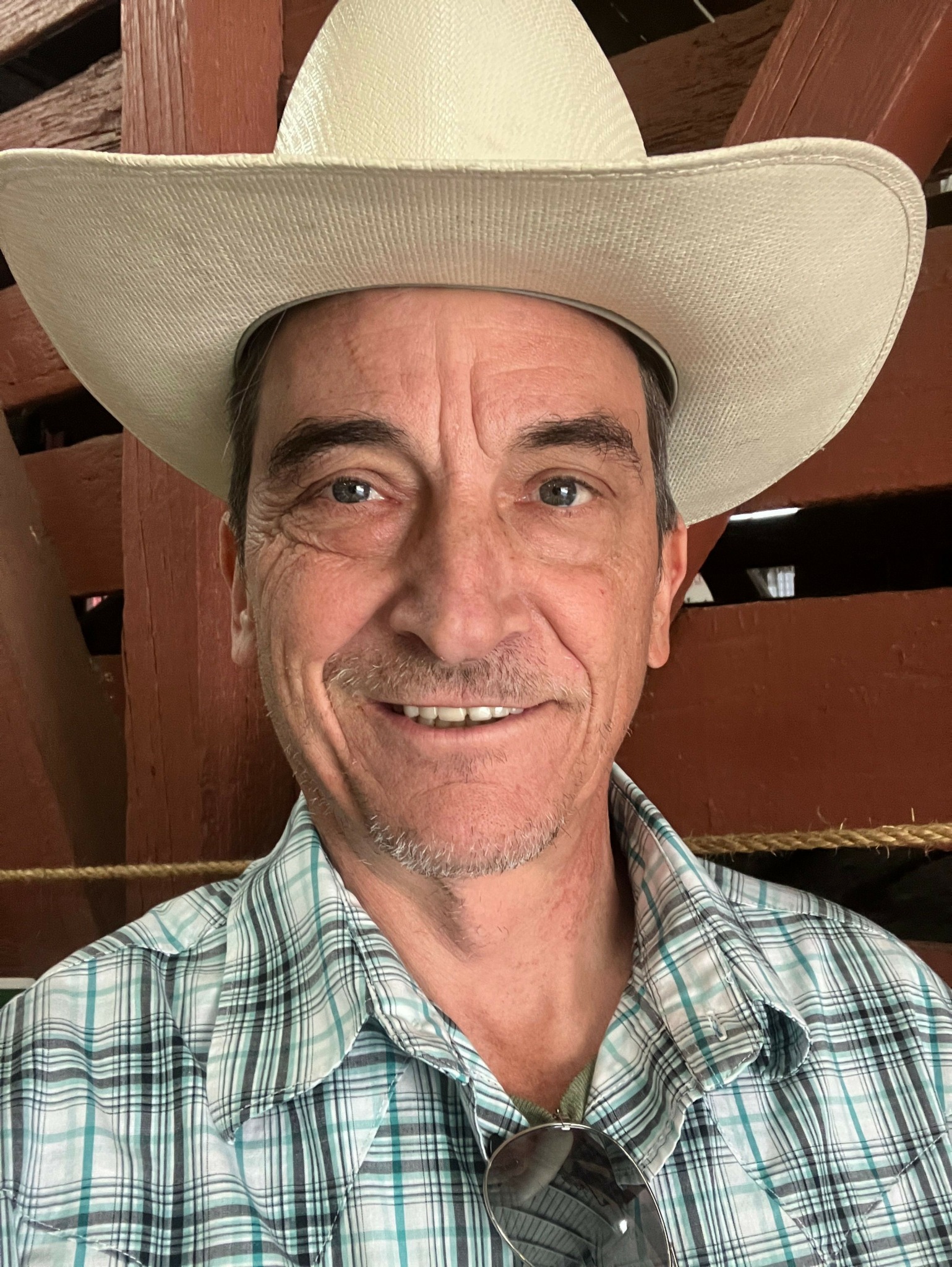 Man wearing a large cowboy hat and plaid shirt, smiling, indoors with wooden furniture in background.