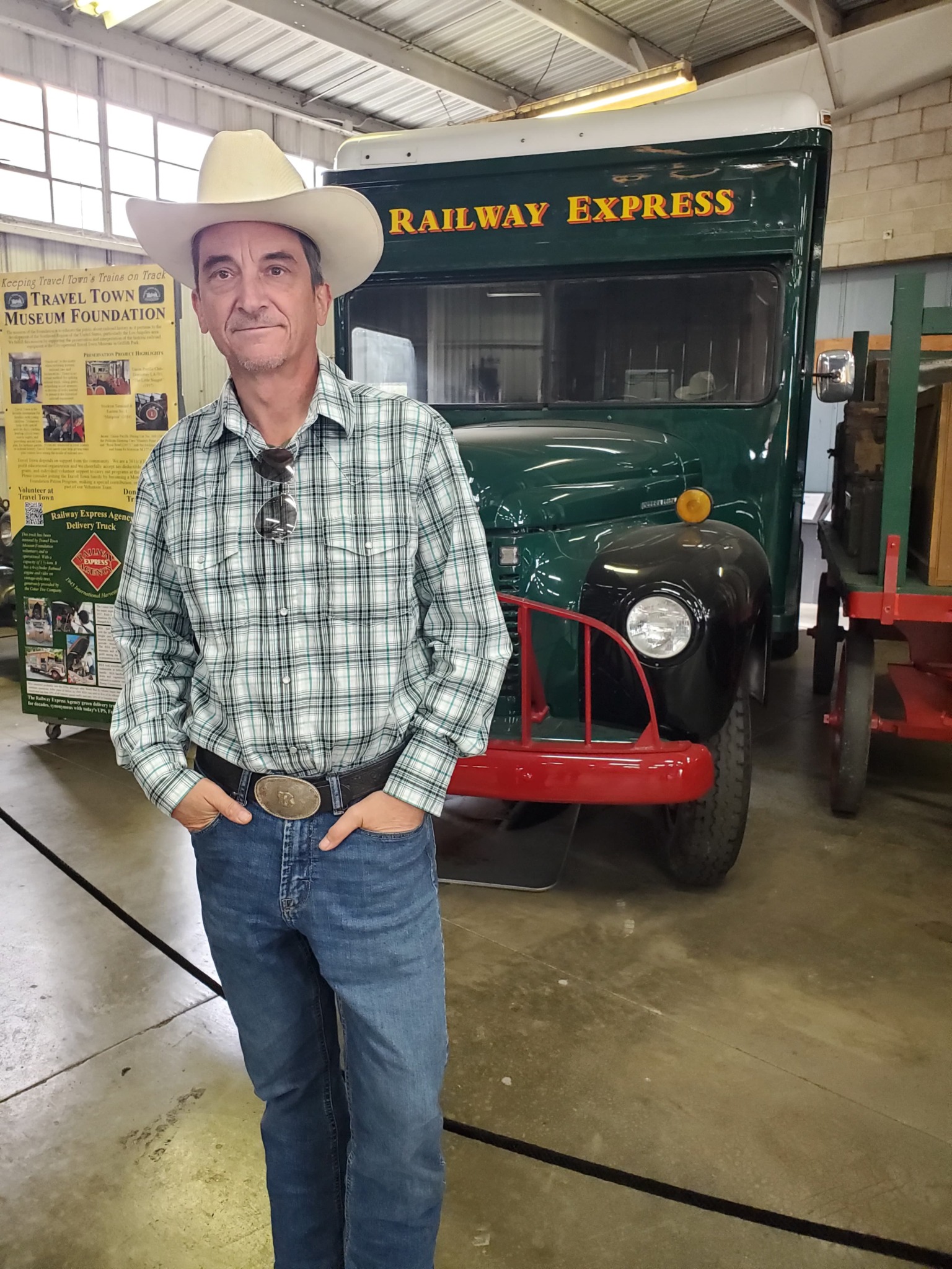 Man in cowboy hat and plaid shirt standing in front of a green vintage train vehicle inside a building.