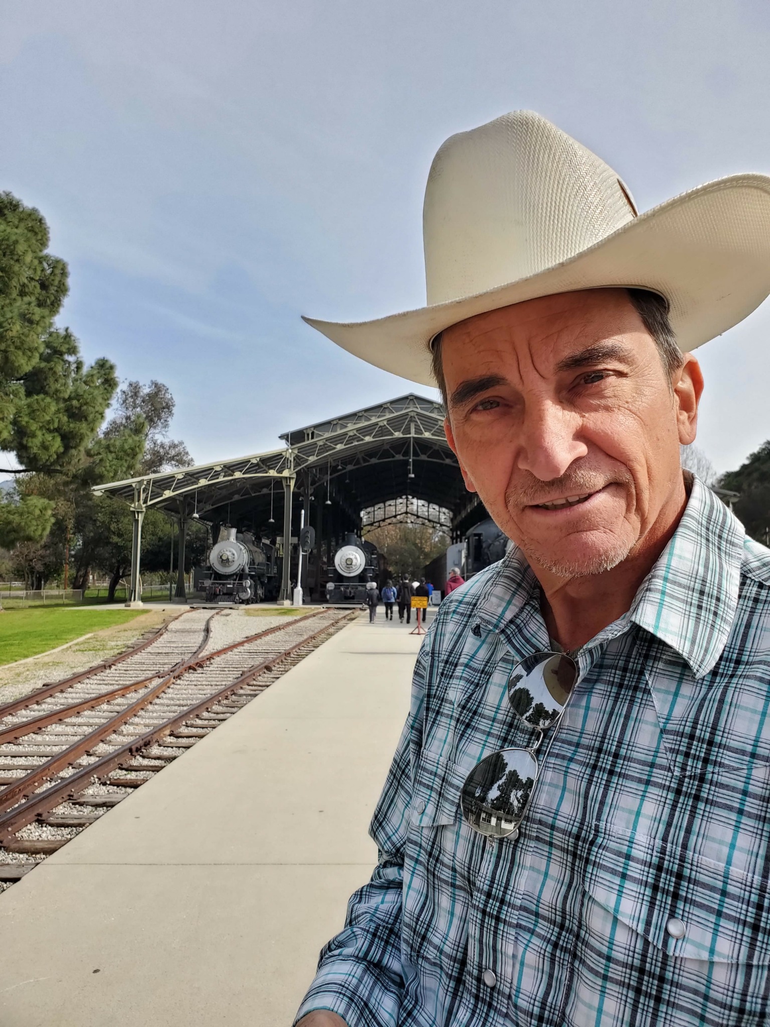 Man wearing a cowboy hat and plaid shirt standing near train tracks outdoors.