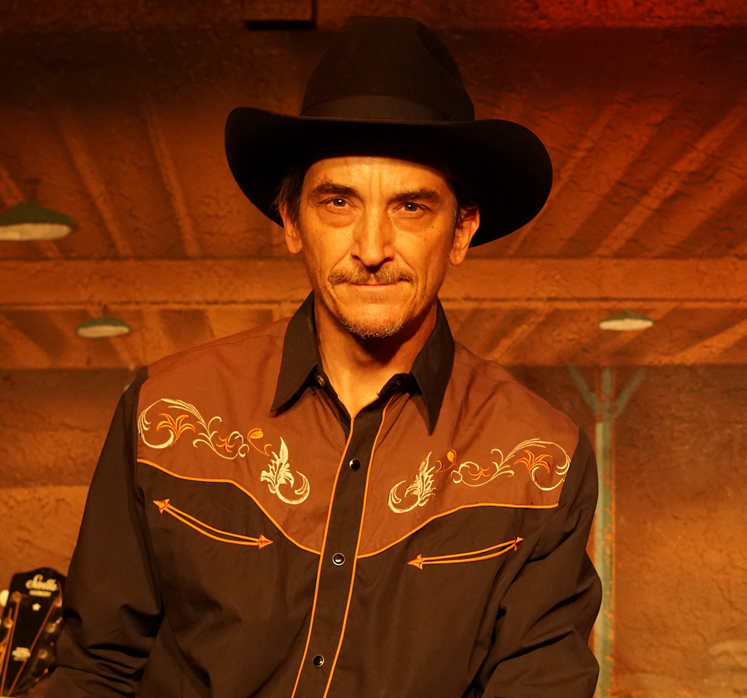 Man wearing a black hat and western-style shirt with embroidery, standing indoors with wooden ceiling.