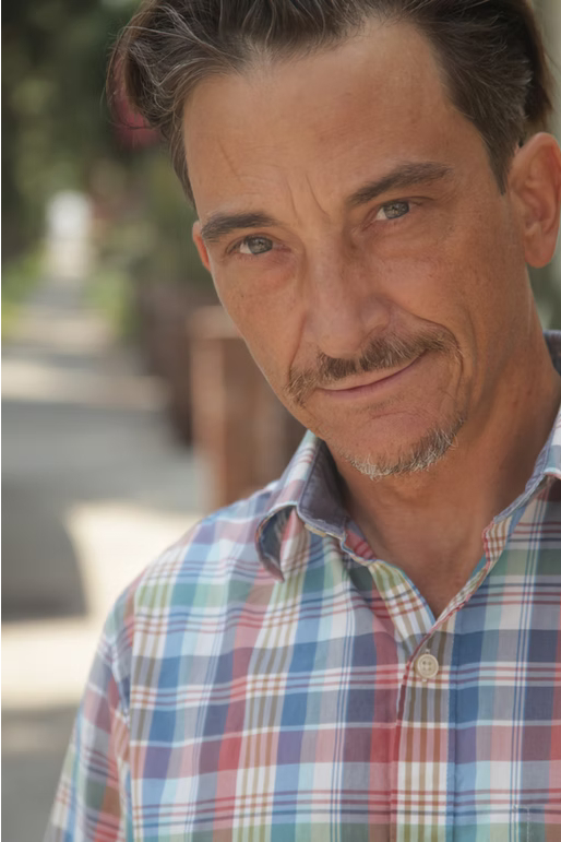 Close-up of a man with short dark hair, wearing a plaid shirt, outdoors with blurred background.