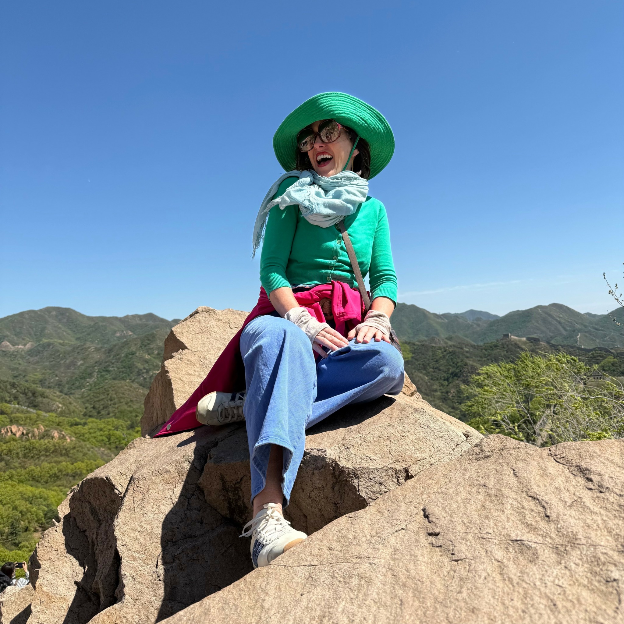 Person sitting on a large rock outdoors, wearing a green hat, glasses, and colorful clothing, with mountains in the background.