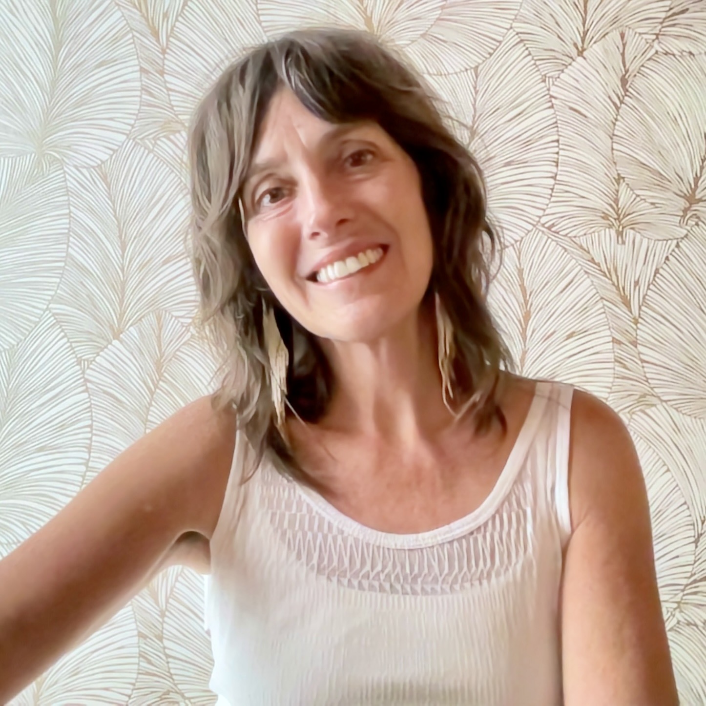 Woman with shoulder-length wavy hair smiling, wearing a sleeveless light-colored top, standing in front of patterned wallpaper.