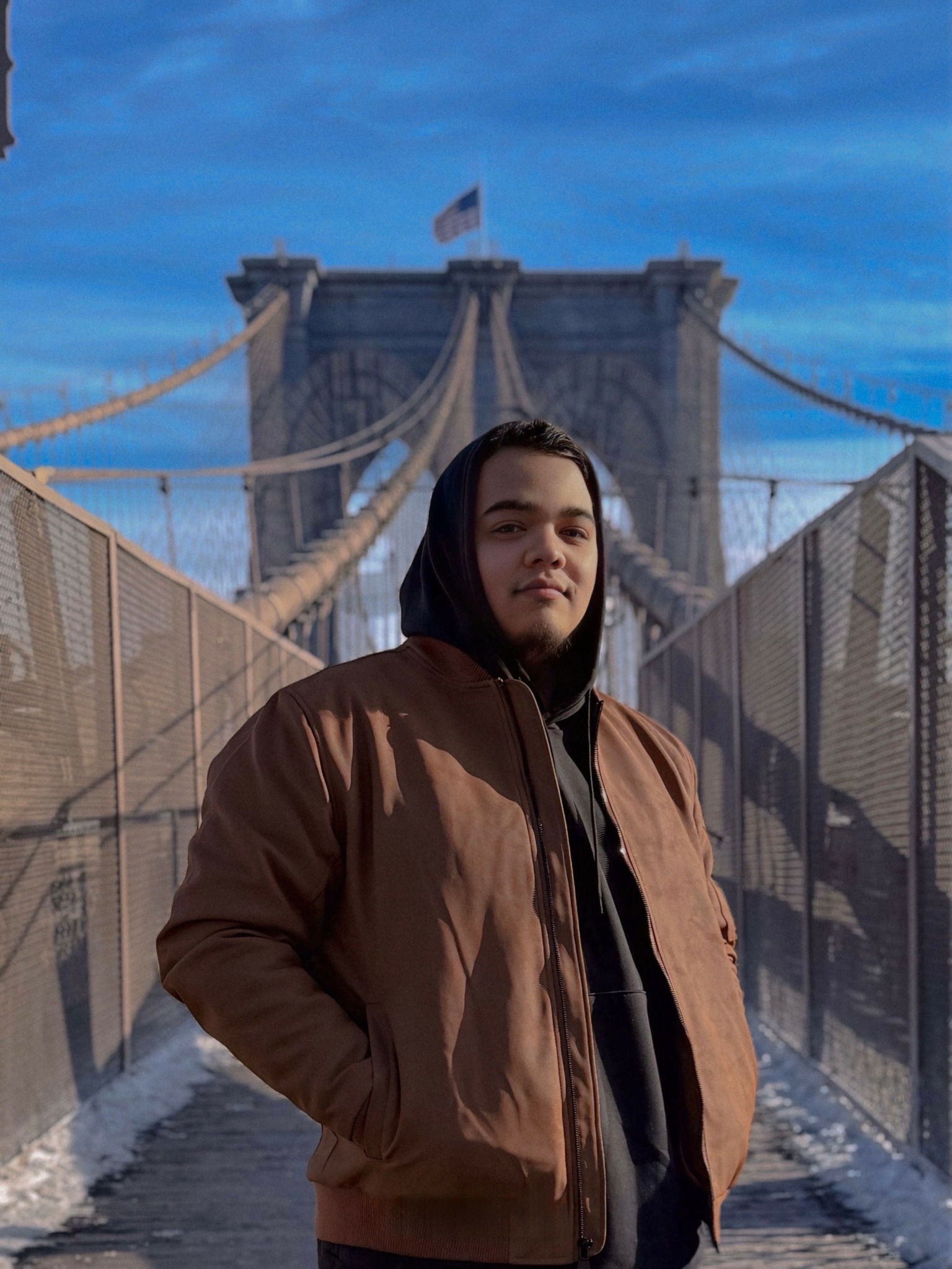 Young person wearing a brown jacket and black hoodie standing on a bridge with a cityscape and bridge structure in background.