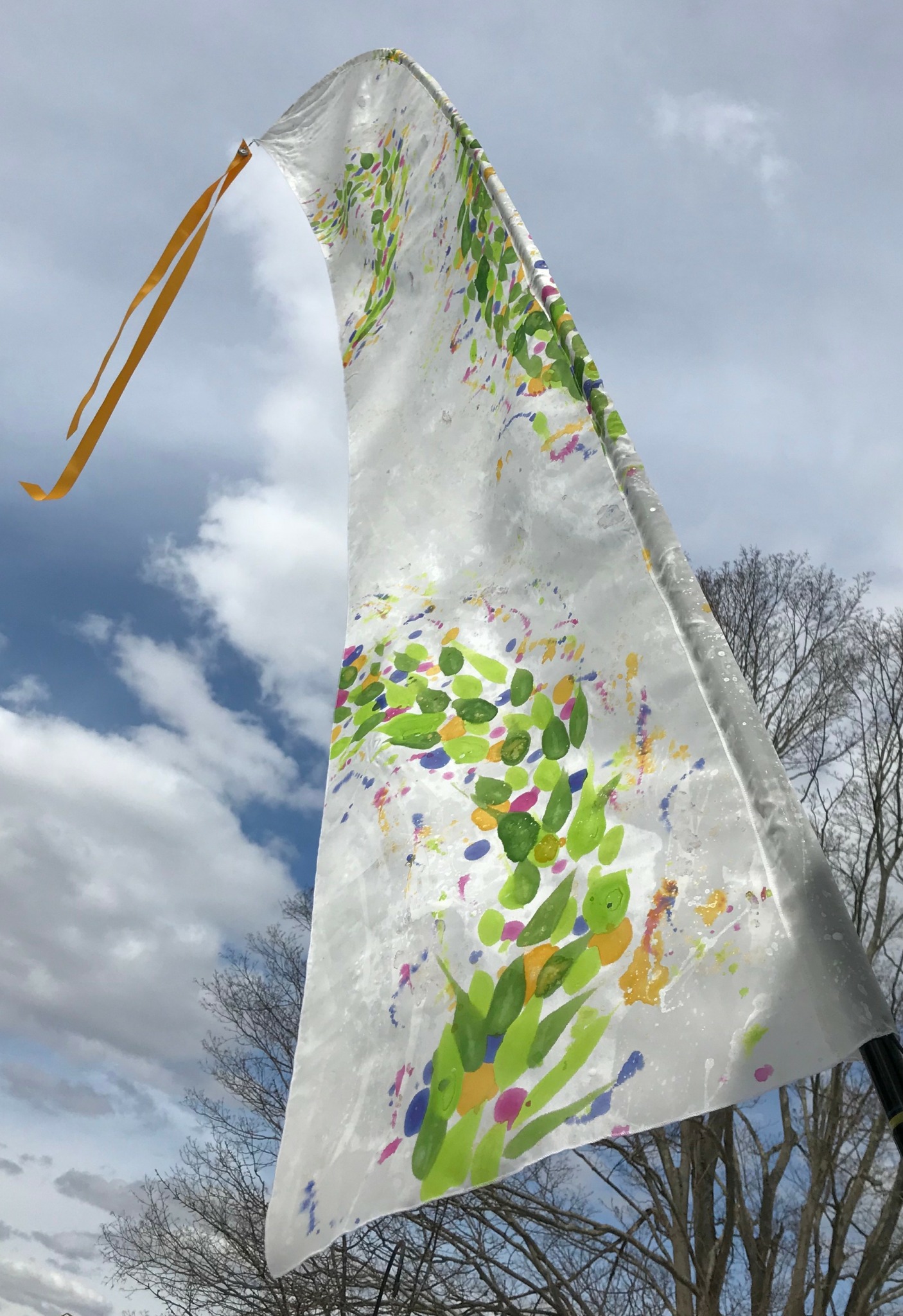 Colorful fabric with green, purple, and yellow patterns waving in the sky, with clouds and trees in the background.