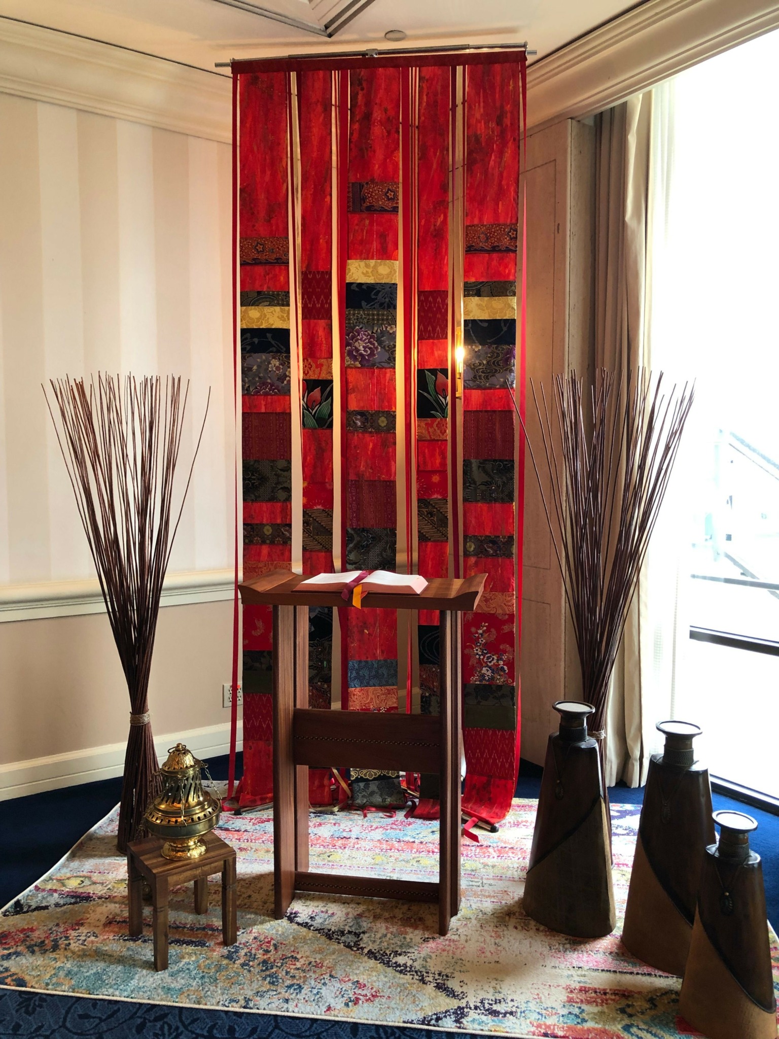 Red fabric hanging panel with gold, black, and red patterns, flanked by tall vases with dried branches, in a room.
