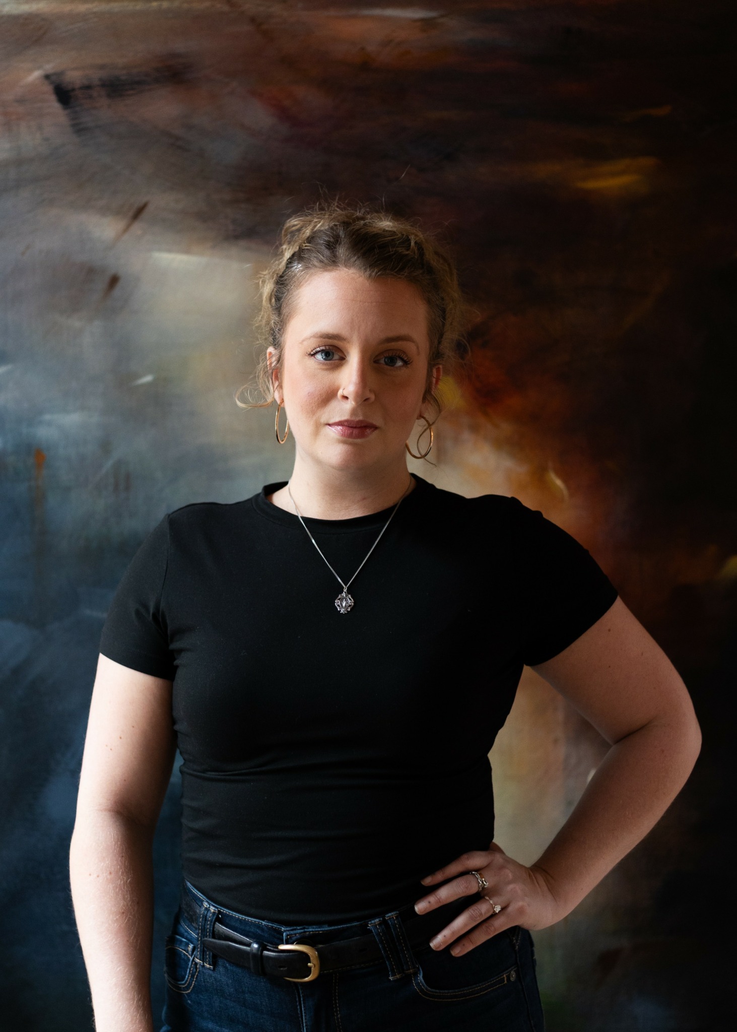Young woman with curly hair in a black t-shirt and jeans, standing with hand on hip against an abstract background.