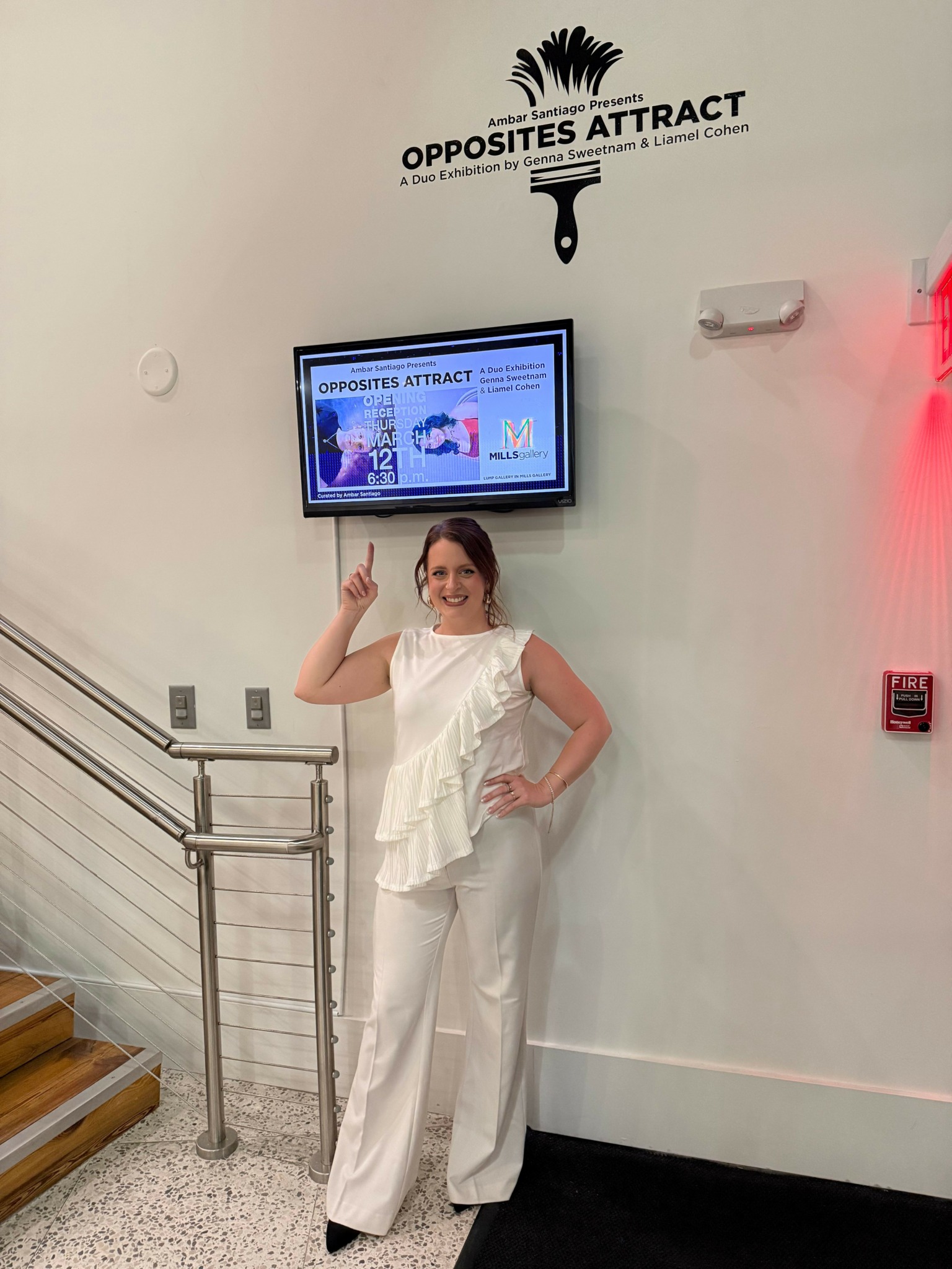 Woman standing next to wall with sign and screen, smiling and pointing upward, in a well-lit indoor space.