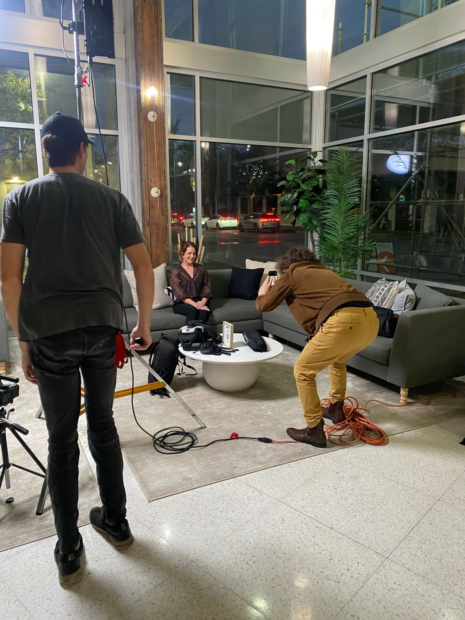 Three people in a modern indoor space with large glass windows, furniture, and plants. One person is seated, two are standing.