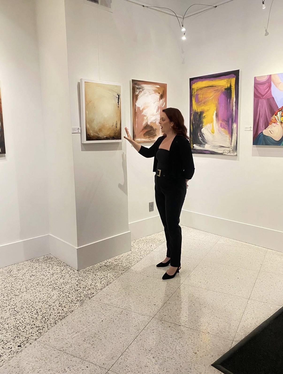 Woman in black pants and blazer looking at abstract paintings in art gallery.