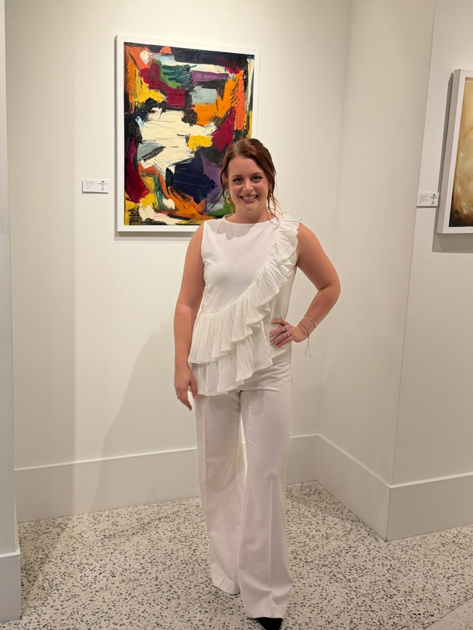 Woman in white outfit standing in art gallery with colorful abstract painting behind her.