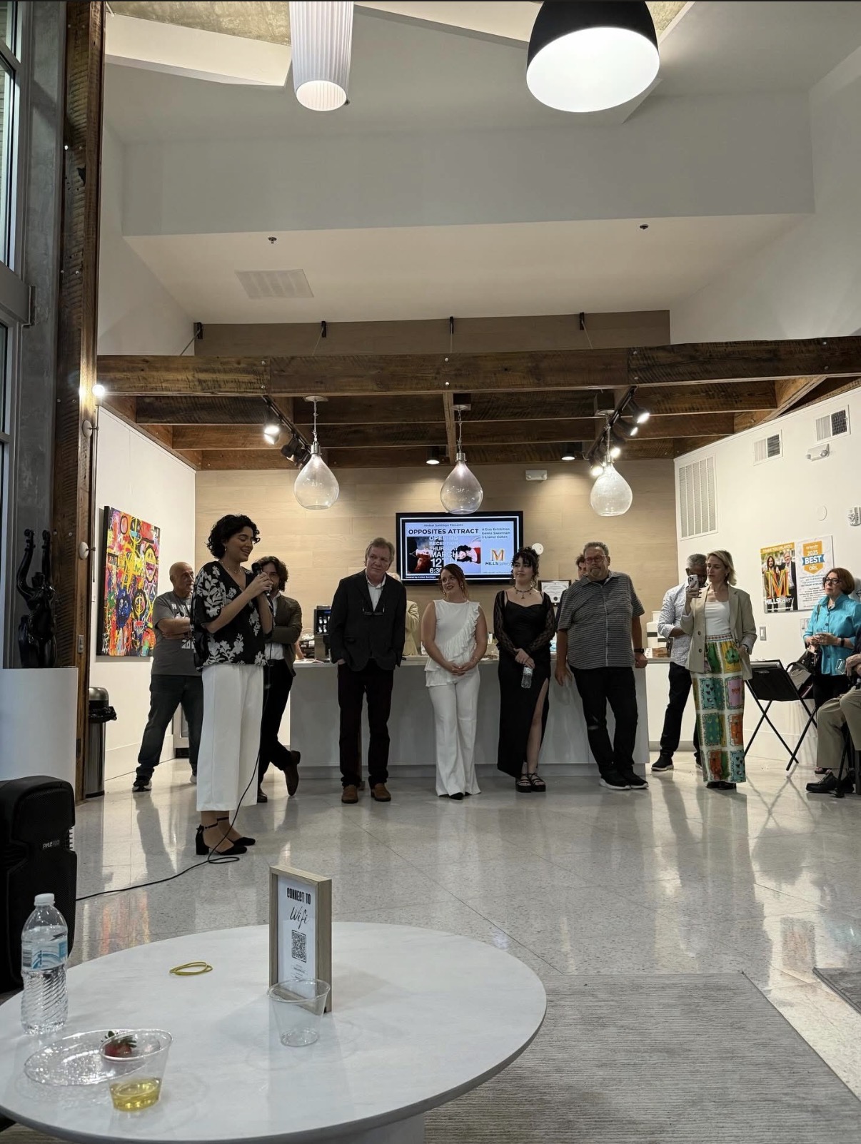 Group of people standing and talking in an art gallery with paintings on walls and a television screen in the background.