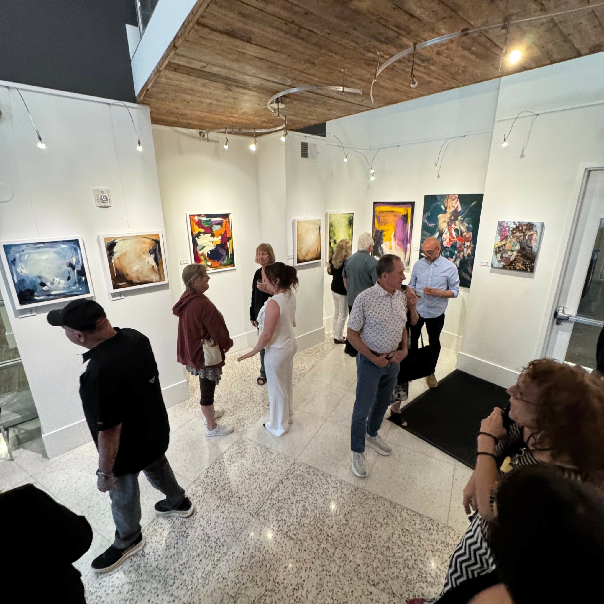 People viewing colorful paintings in an art gallery with white walls and wooden ceiling.