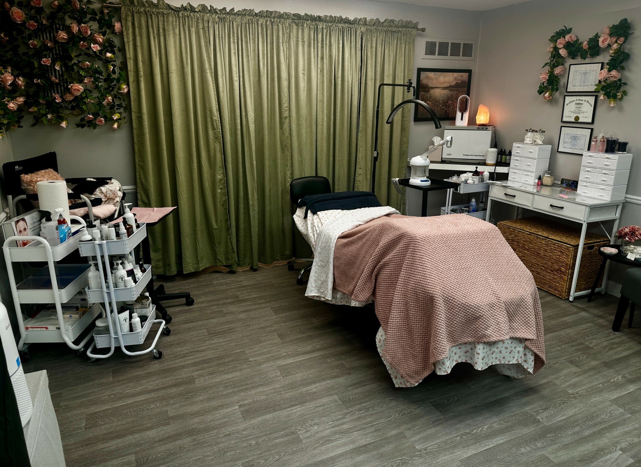 Room with massage table, green curtains, shelves with boxes, and decorative flowers on wall.