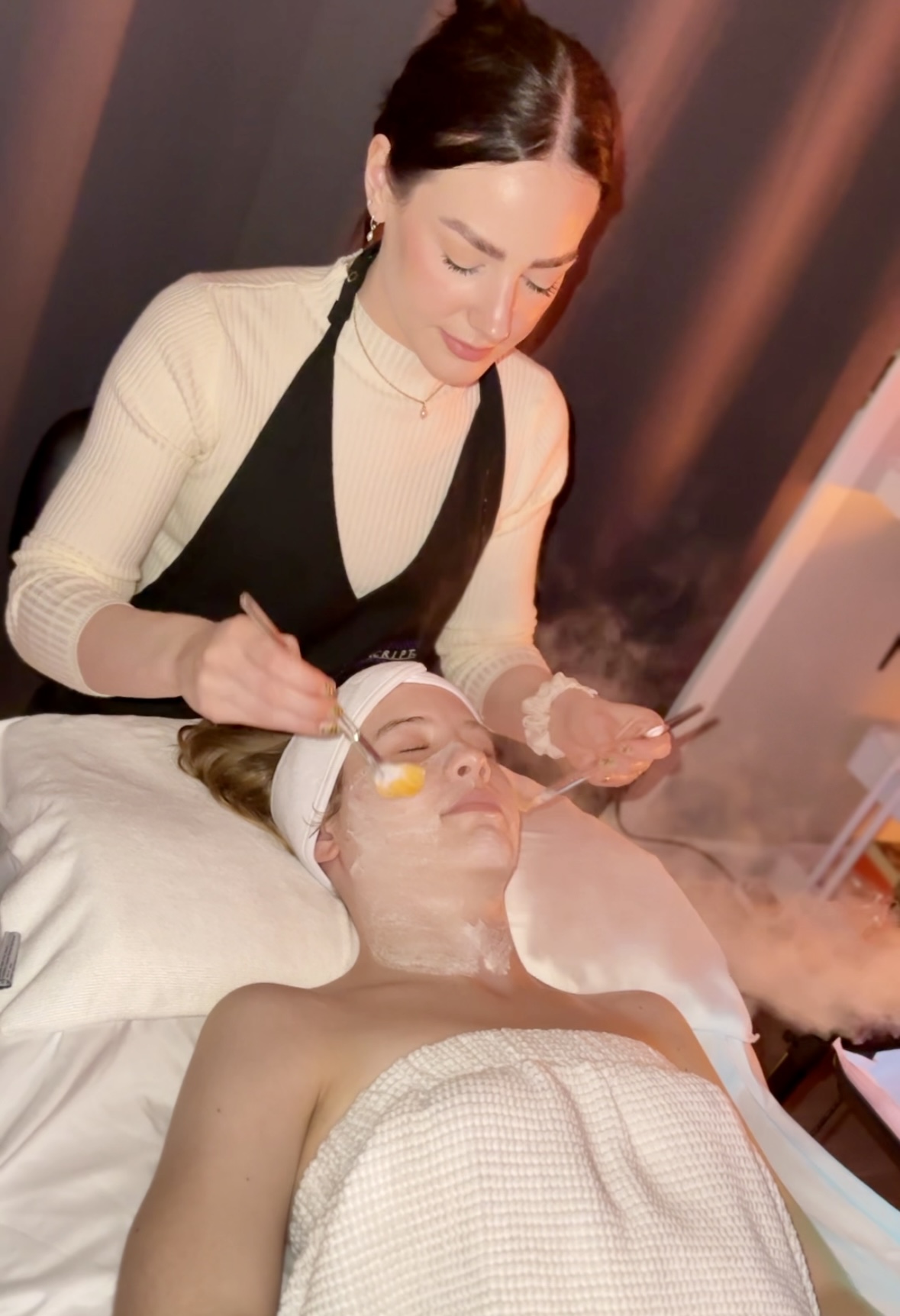 A woman in a black and beige outfit performs a facial treatment on a person lying in a spa bed.