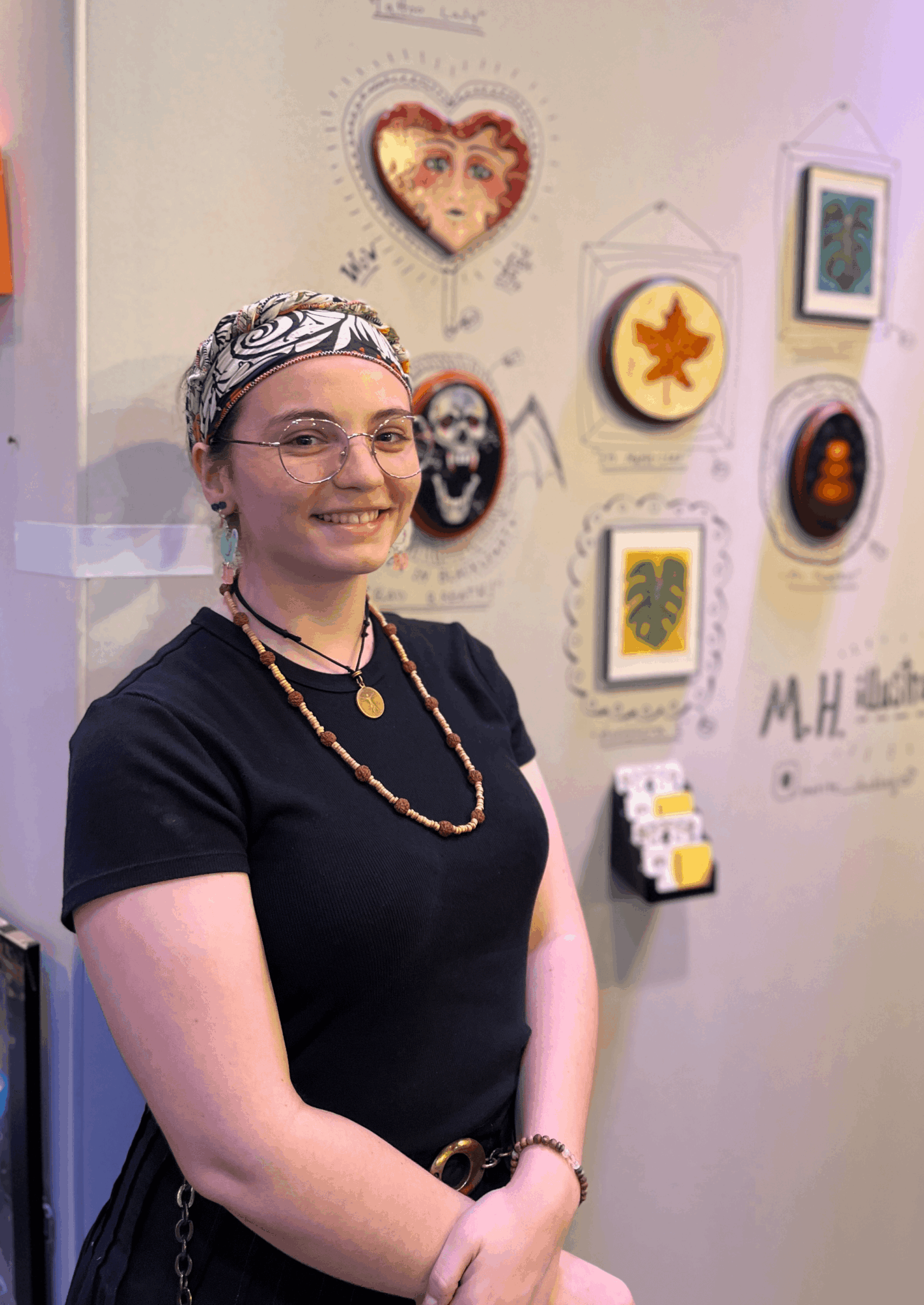Smiling woman with glasses, wearing a headband and jewelry, standing in front of art pieces on a wall.