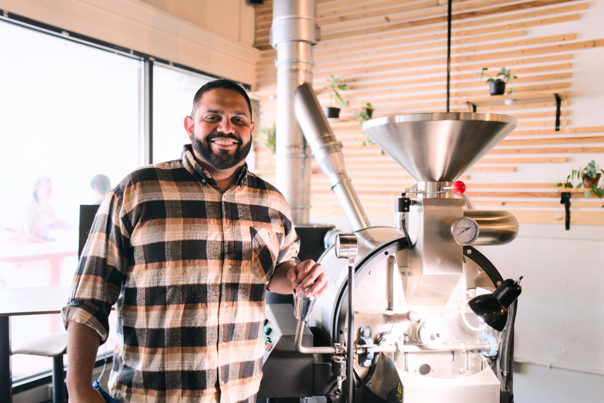 Man with a beard in a plaid shirt standing next to a coffee roasting machine inside a cafe.