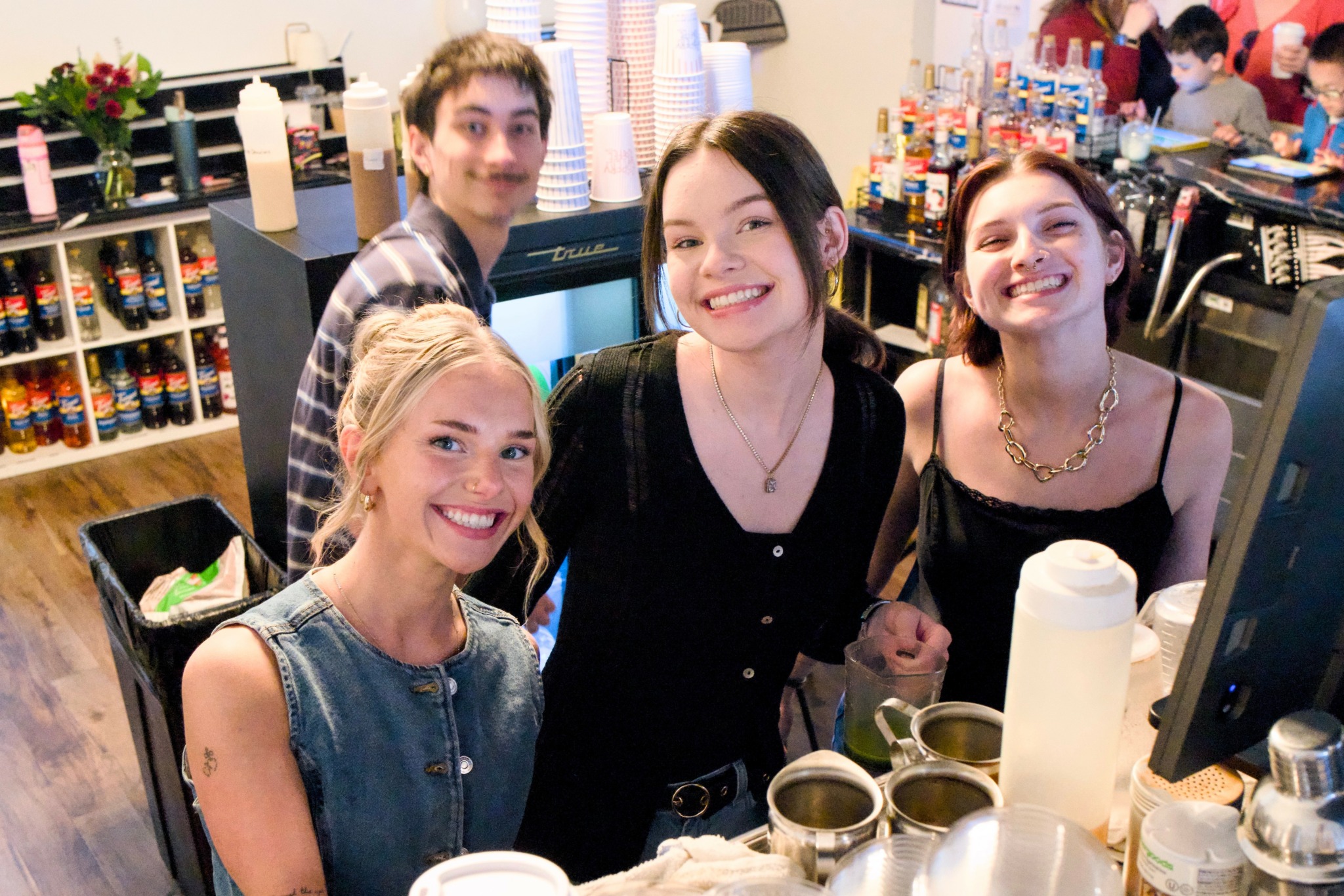 Four smiling young women at a bar or cafe counter with drinks and a computer screen.