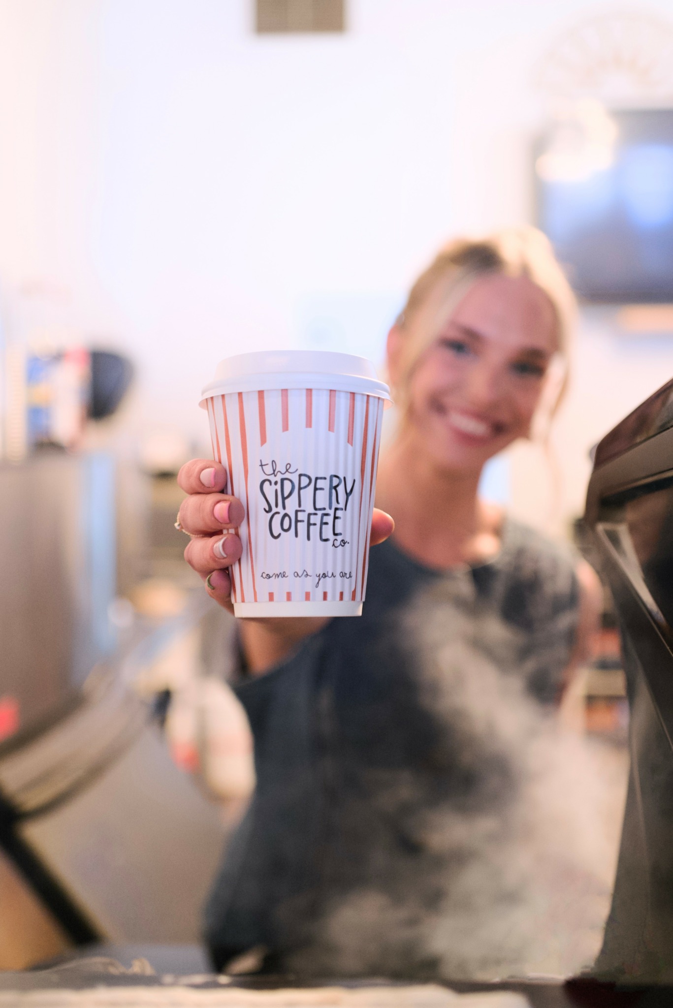 Person holding a coffee cup with striped design and text, smiling in a cafe setting.