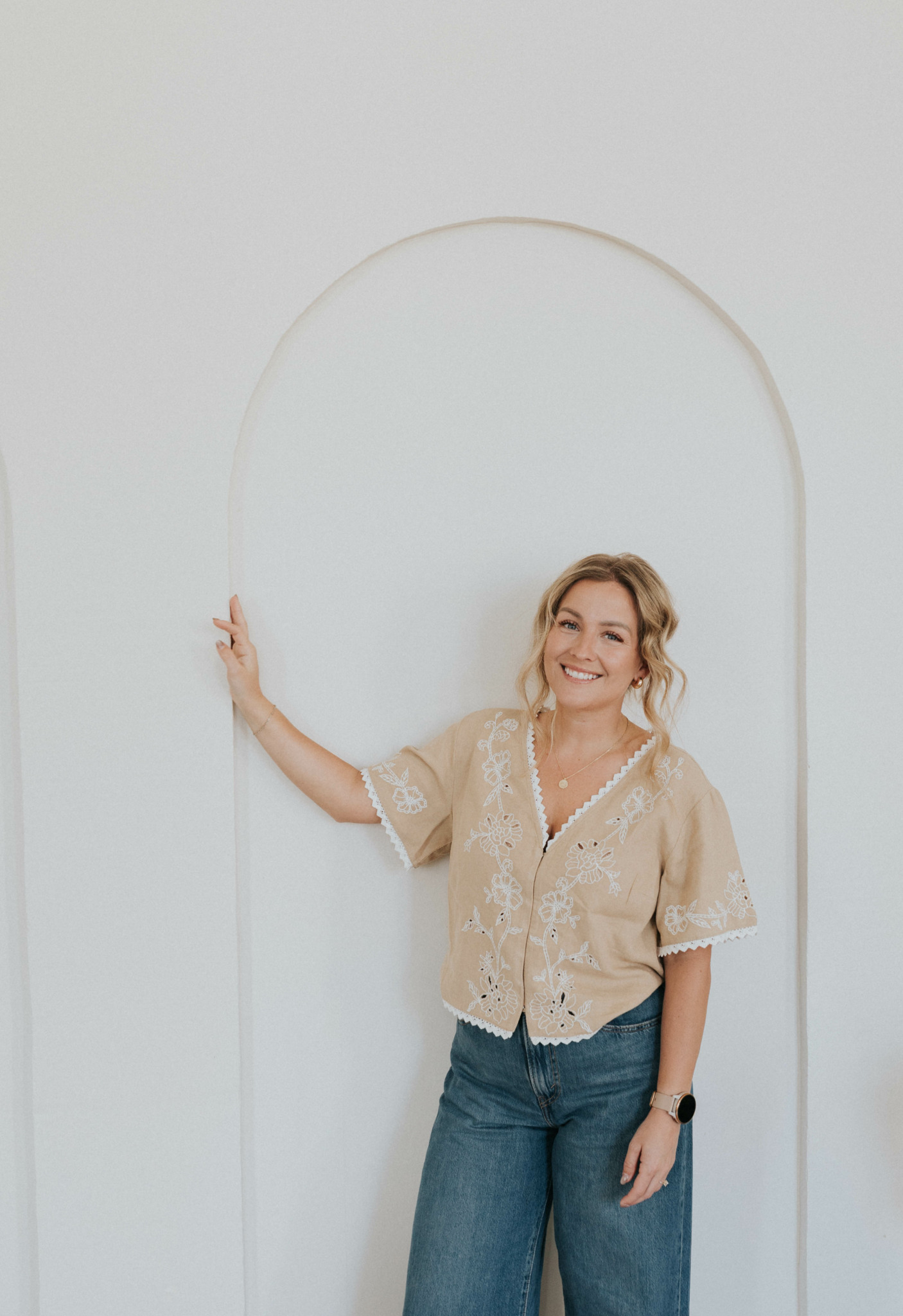 Smiling woman with blonde hair in a beige blouse and jeans, standing next to a white wall with an arch.