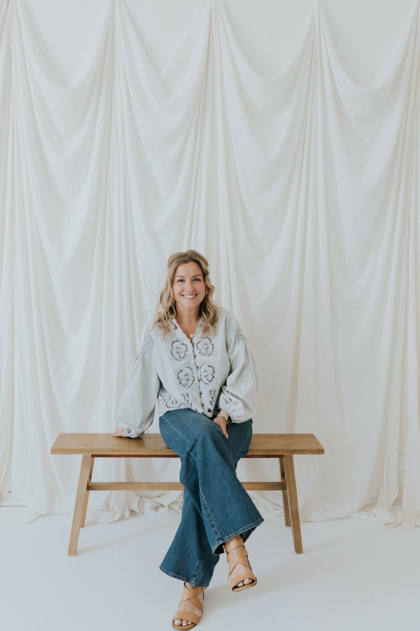 Woman sitting on a wooden bench in front of a white curtain backdrop, smiling, wearing a white blouse and blue jeans.