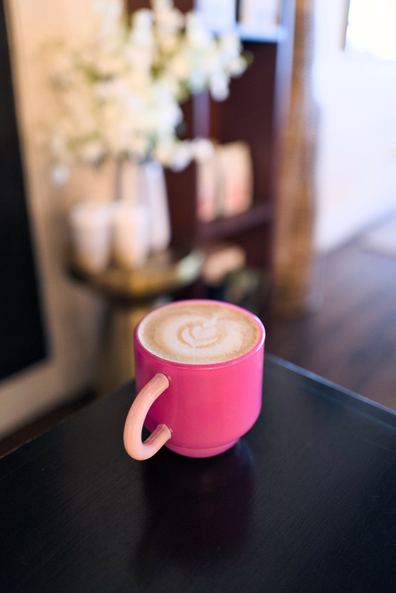 Pink mug with latte art on a black table, blurred background with flowers and furniture.