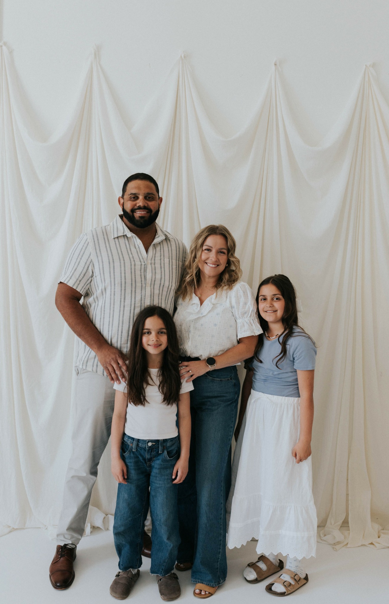 Four people stand in front of a white draped backdrop, smiling, with two adults and two children.