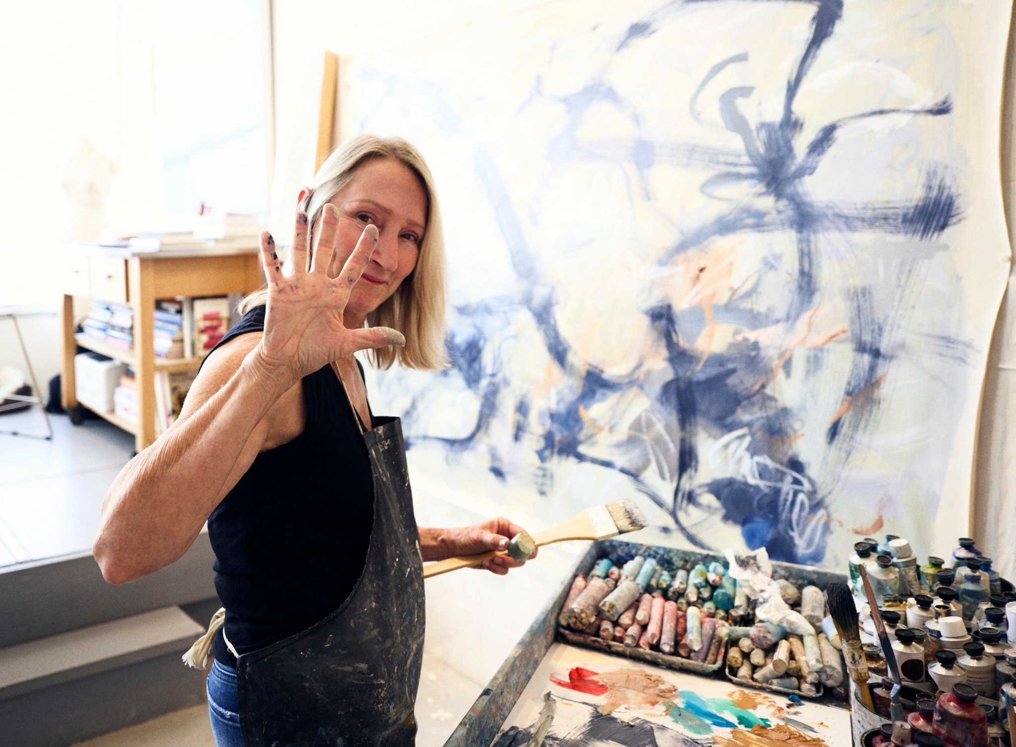 Woman in a black top and apron raises her hand near a large abstract painting on the wall.
