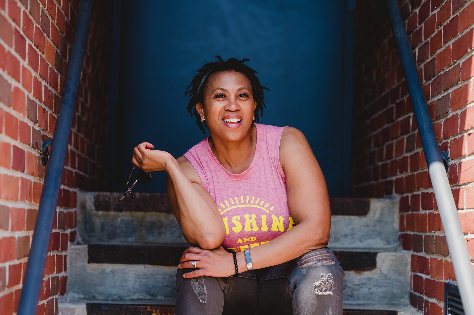 Smiling woman sitting on stairs between brick walls, wearing a pink sleeveless top, holding a bag, with a blue background.
