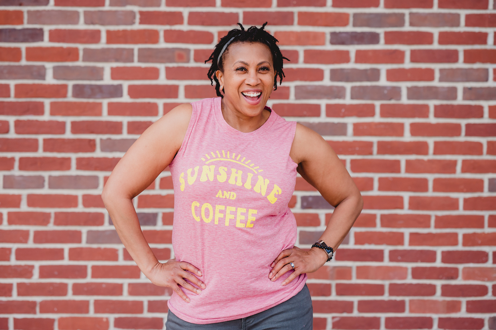 Woman smiling, wearing pink sleeveless shirt with yellow text, standing in front of a brick wall.