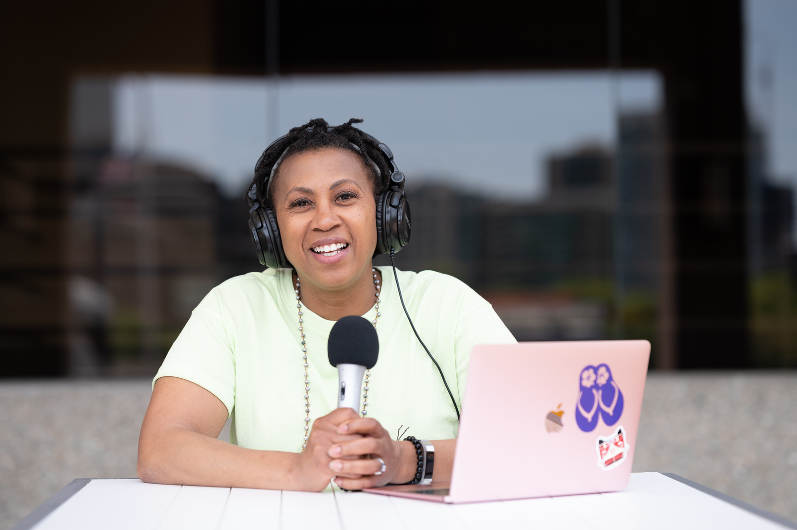 Woman wearing headphones speaking into a microphone at a table with a laptop, outdoors with buildings in background.