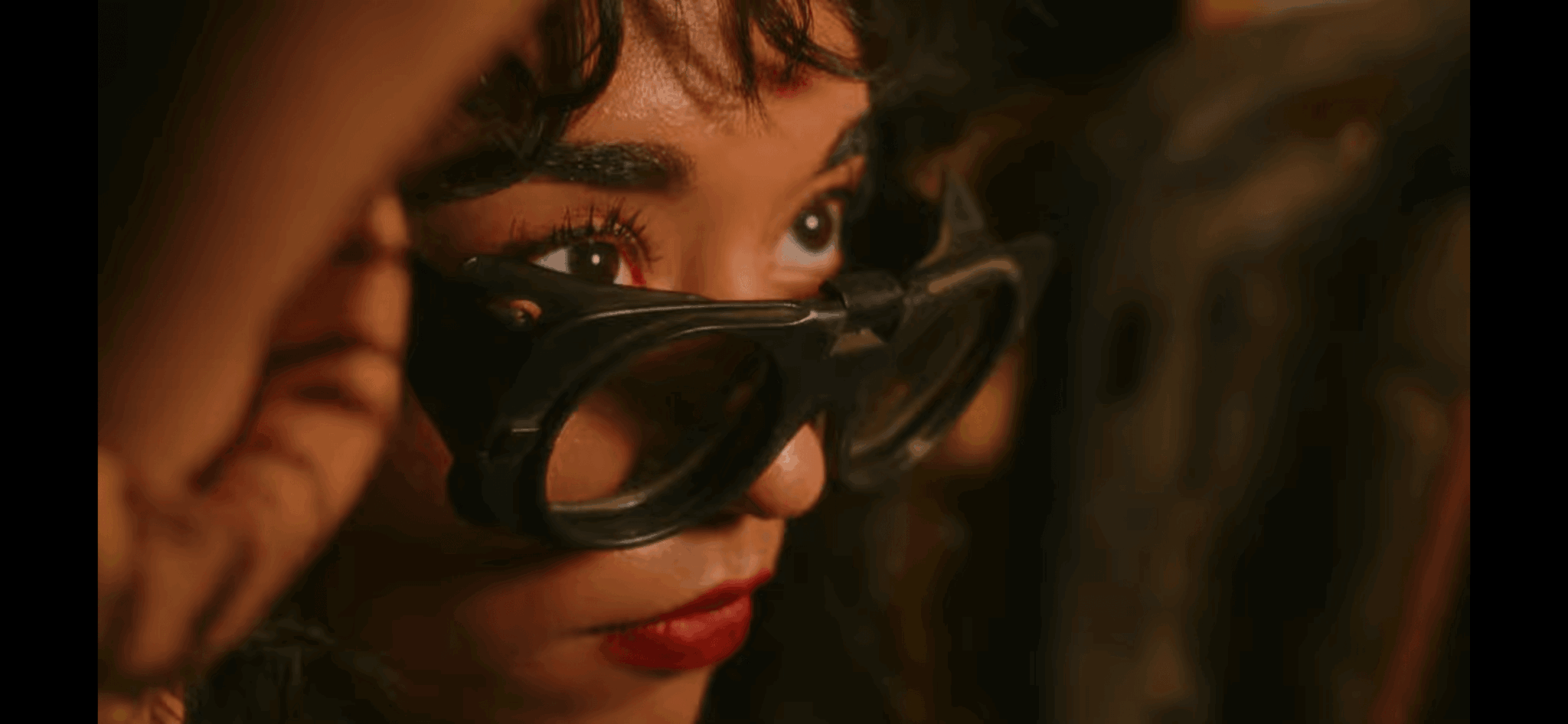 Close-up of a person with glasses, looking intently, with dark curly hair and makeup, in a dimly lit setting.
