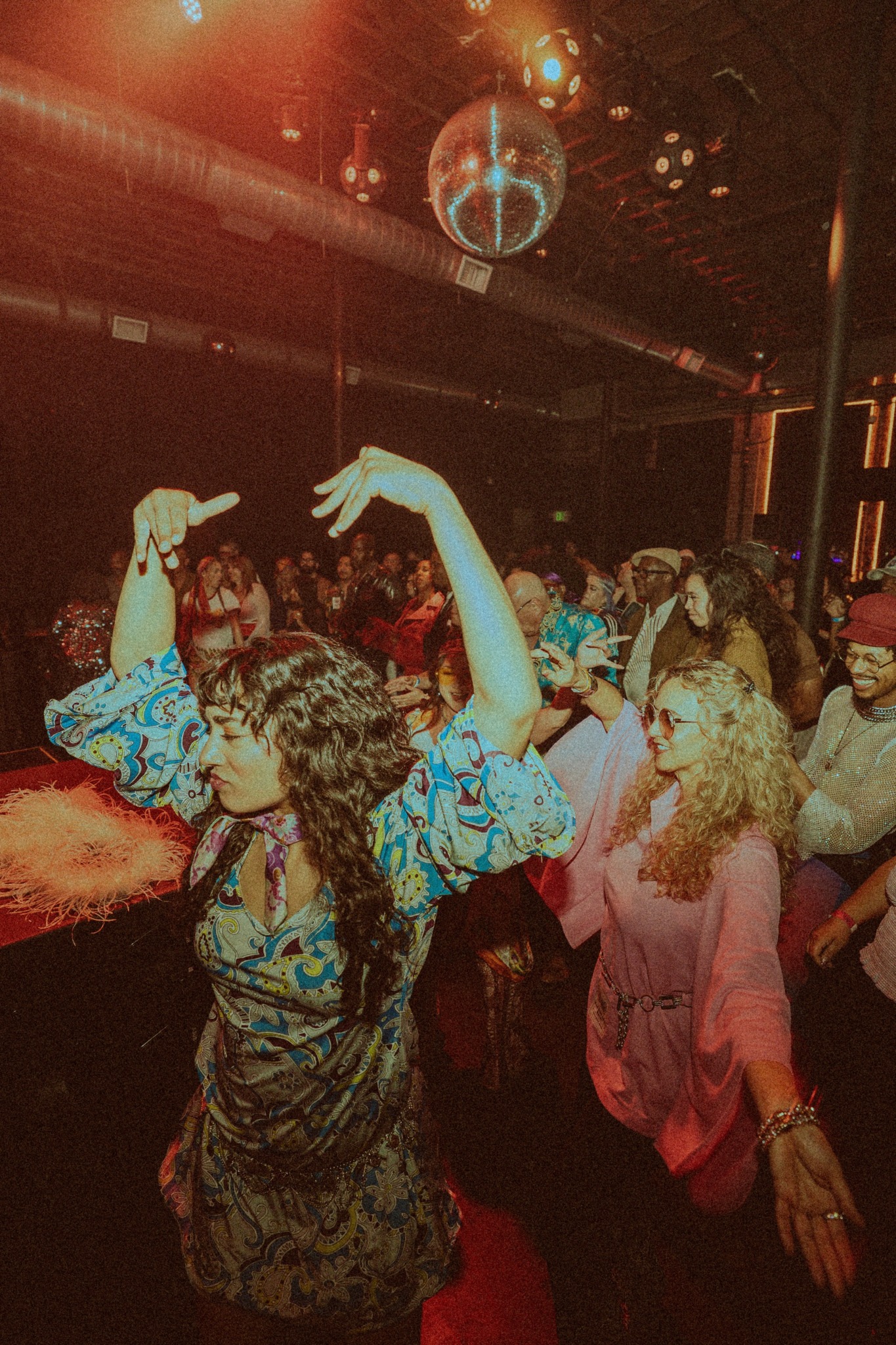 People dancing in a nightclub with colorful lighting and a disco ball overhead.
