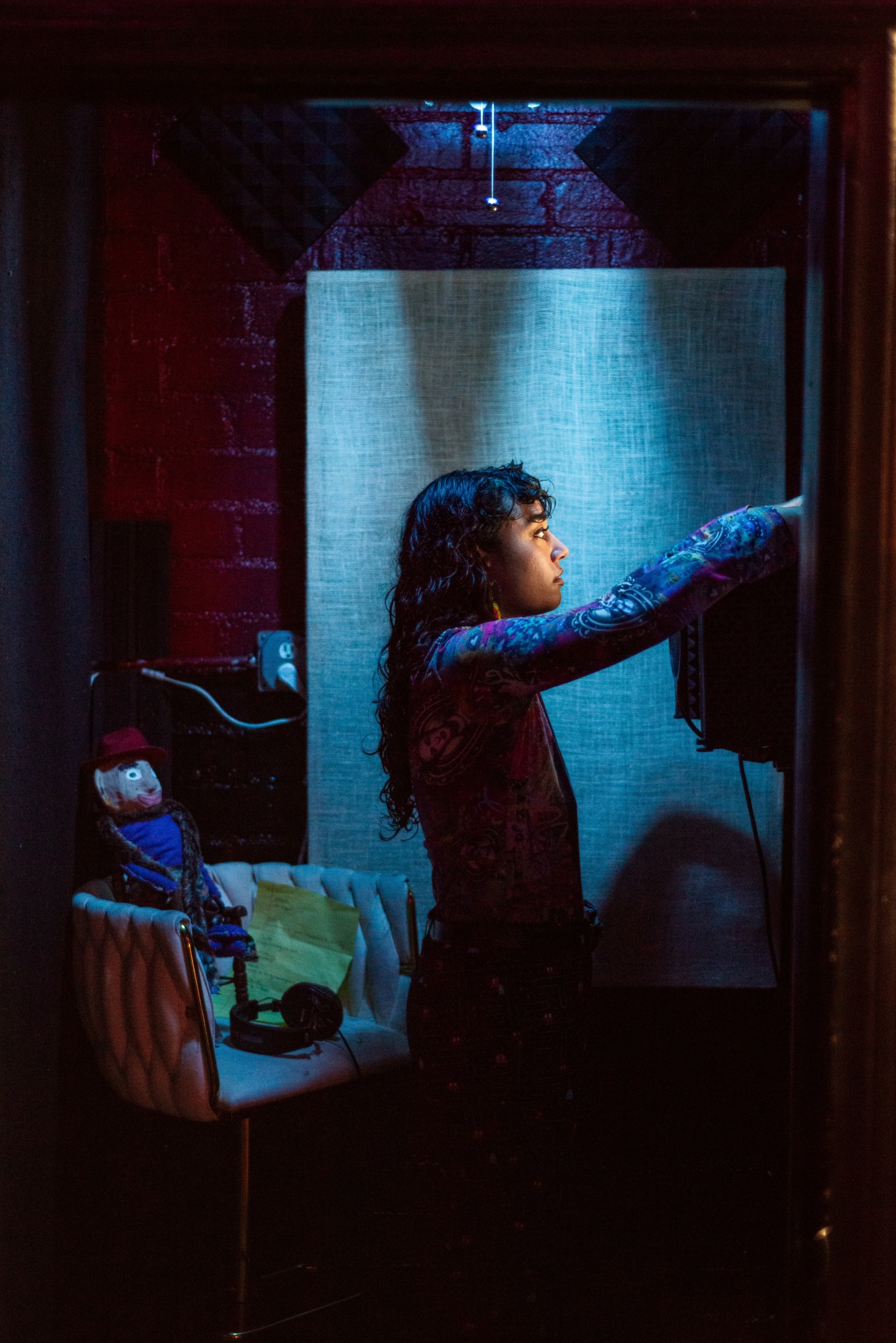 Young girl reaching towards a wall with a dark background and a chair with a doll on it.
