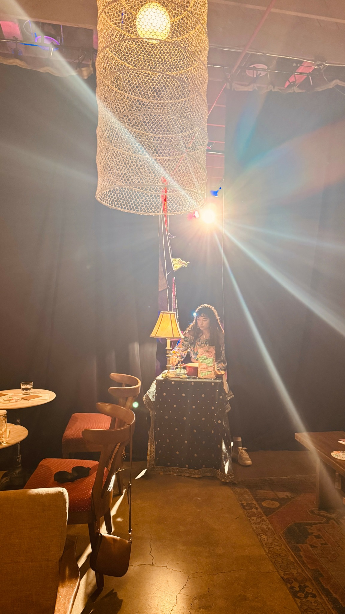 Person standing behind a table with a lamp, surrounded by chairs in a dimly lit room with hanging decorations.