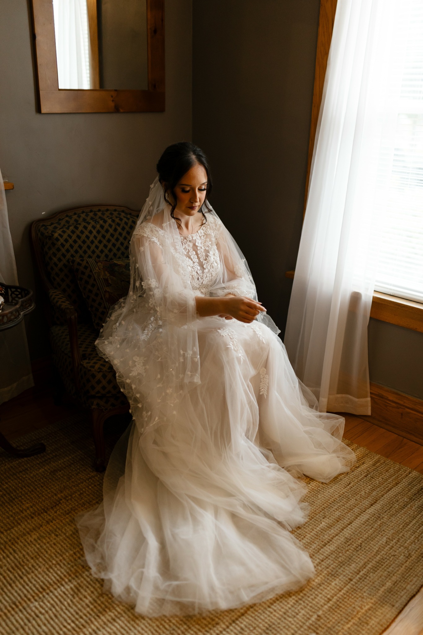 Woman in wedding dress sitting on a chair near window with curtains, holding her hands on her lap, in a room with wooden floor.