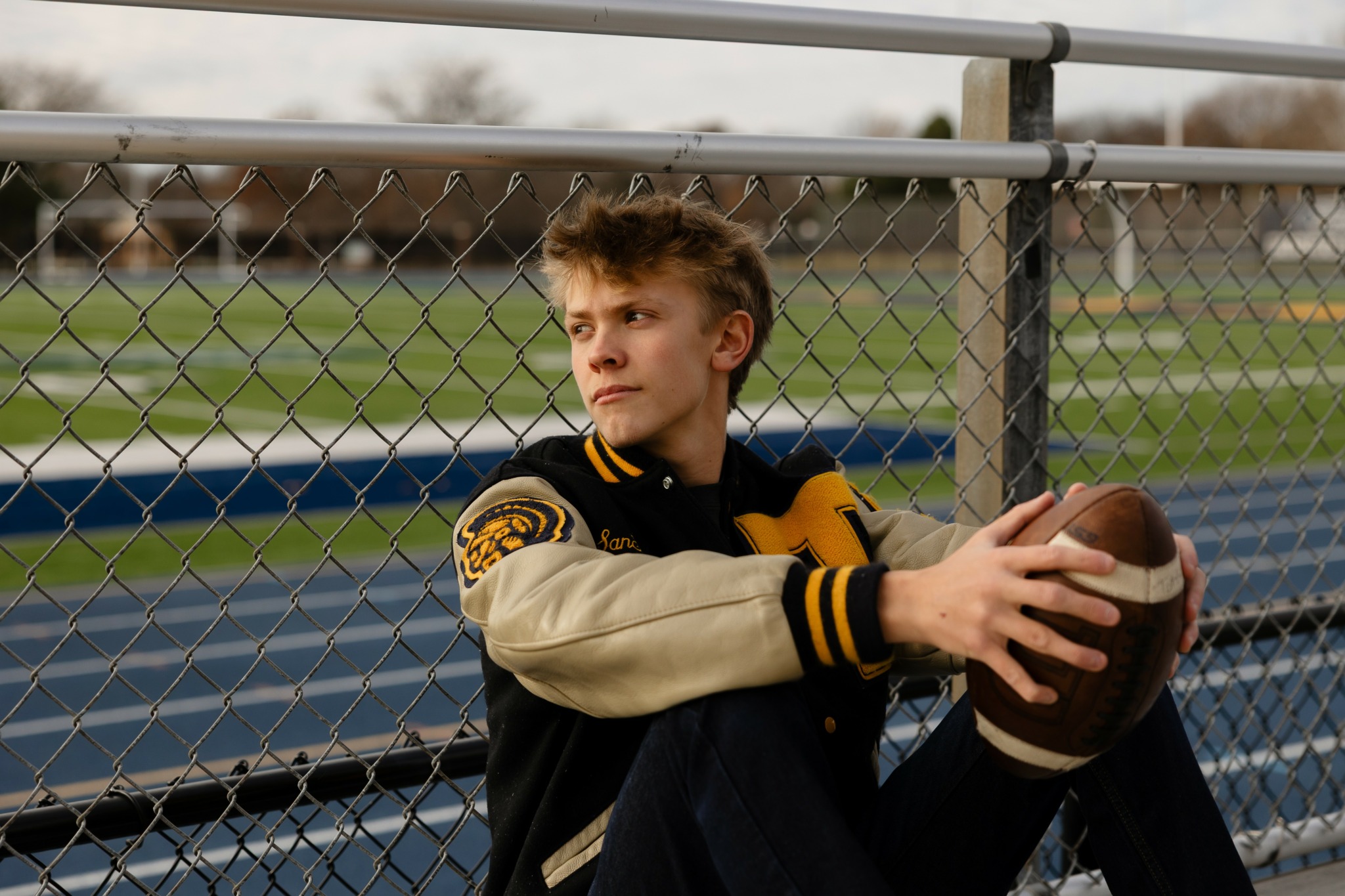 Teenager sitting by a chain-link fence holding a football, wearing a varsity jacket, outdoors on a sports field.
