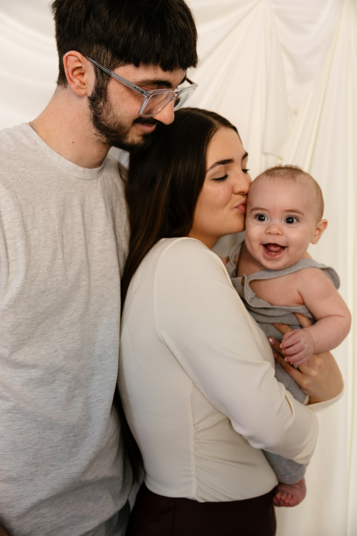 Family of three with a man, woman, and baby, indoors with light-colored drapery background.