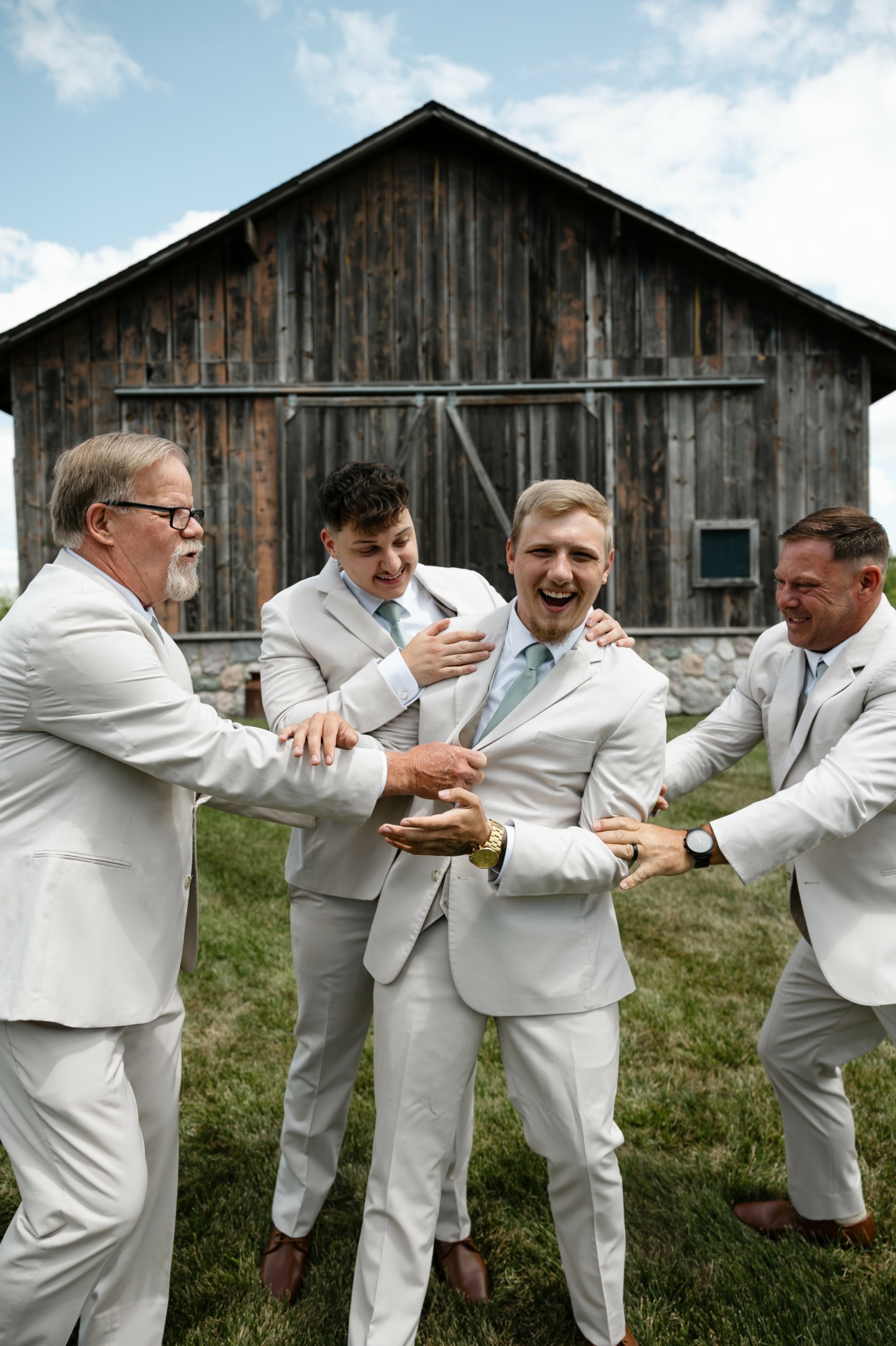 Four men in white suits celebrate outdoors in front of a wooden barn, smiling and holding a trophy.