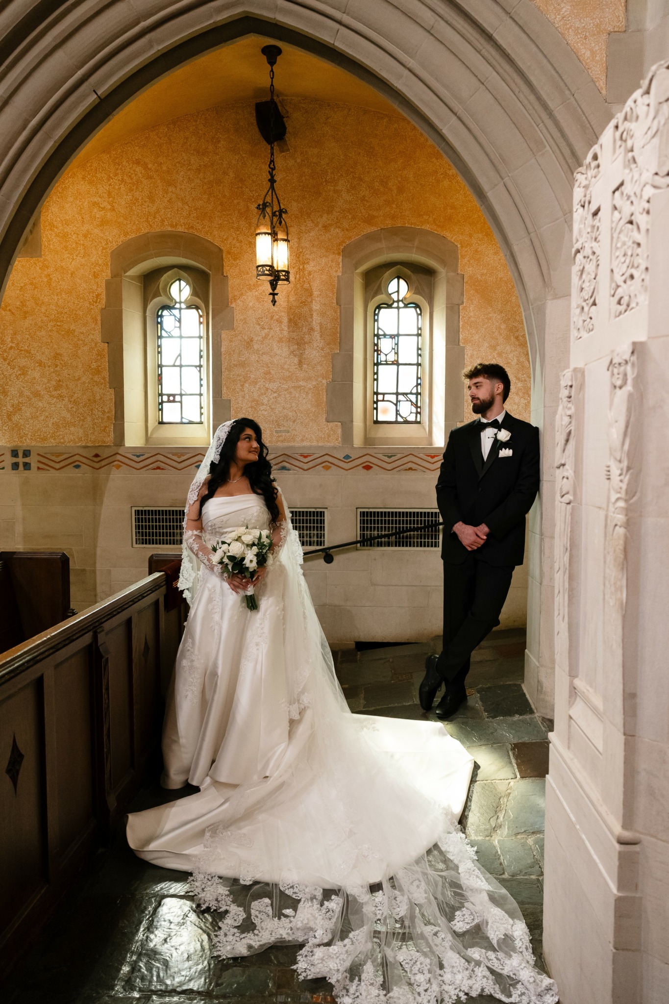 Bride and groom standing inside a church, bride holding a bouquet, church interior with stained glass windows and hanging light.