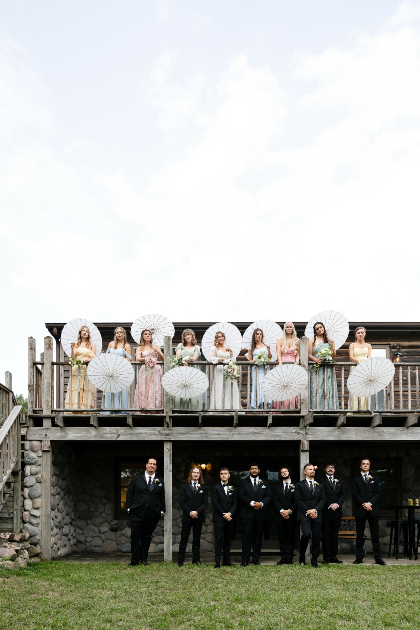 Group of people on a balcony holding umbrellas, with a group of men in suits standing on grass below.