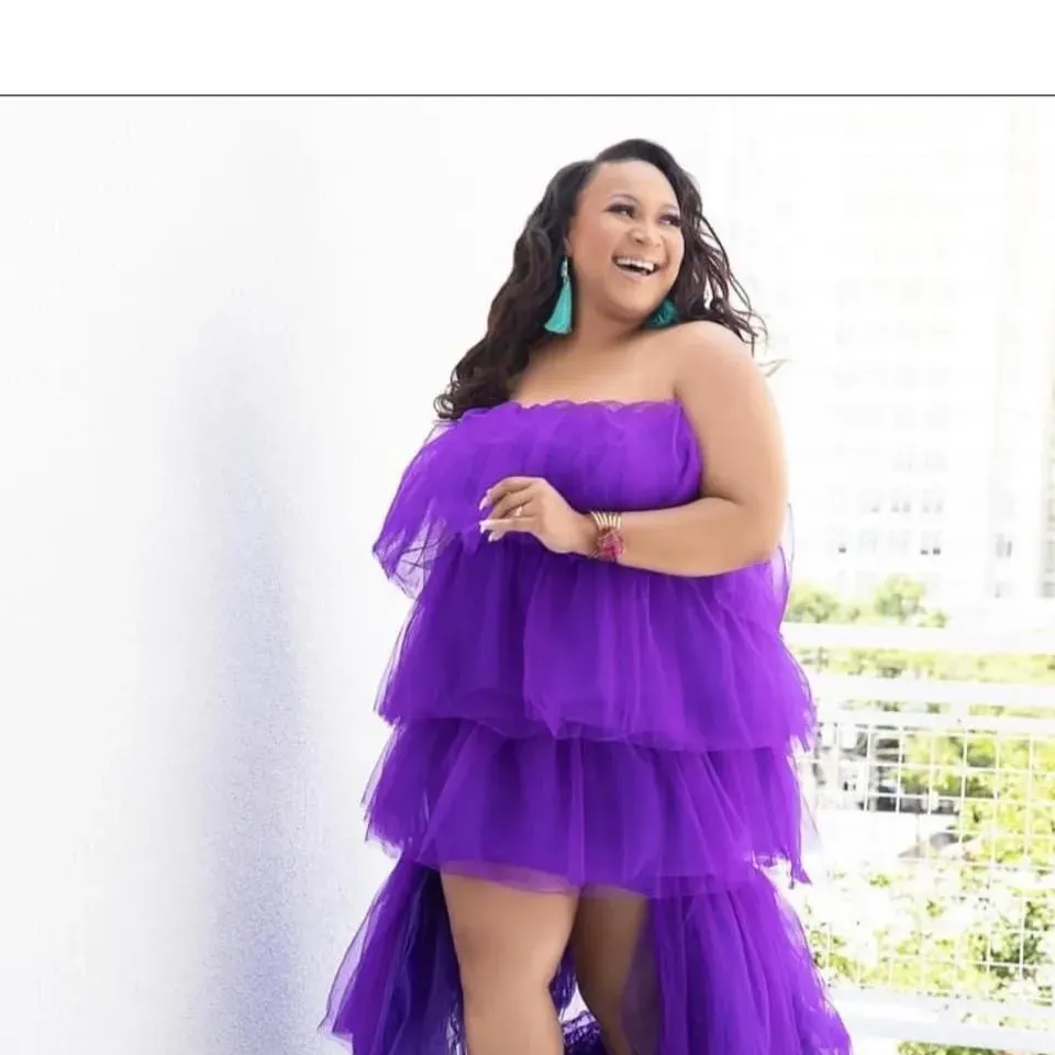 Woman in purple dress smiling outdoors with a balcony and cityscape in background.