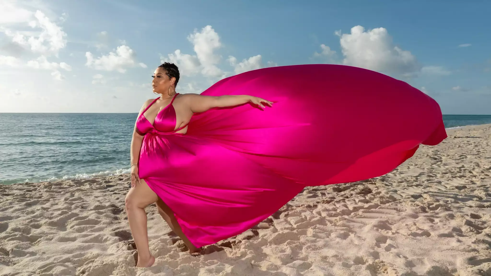 Woman in a pink dress standing on the beach with fabric flowing in the wind, ocean and sky in background.