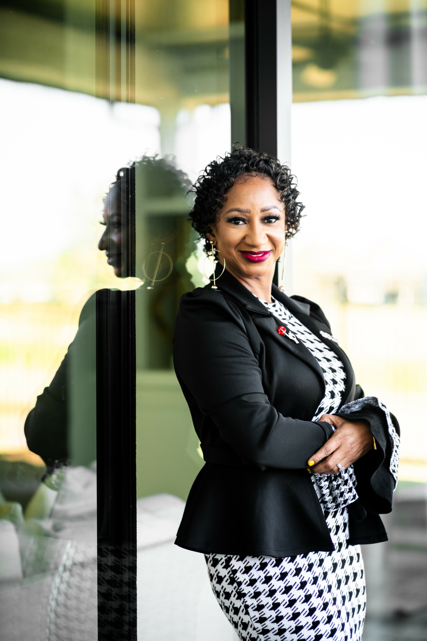 Woman with curly hair and red lipstick standing outdoors near glass wall, smiling, wearing a black blazer and patterned dress.