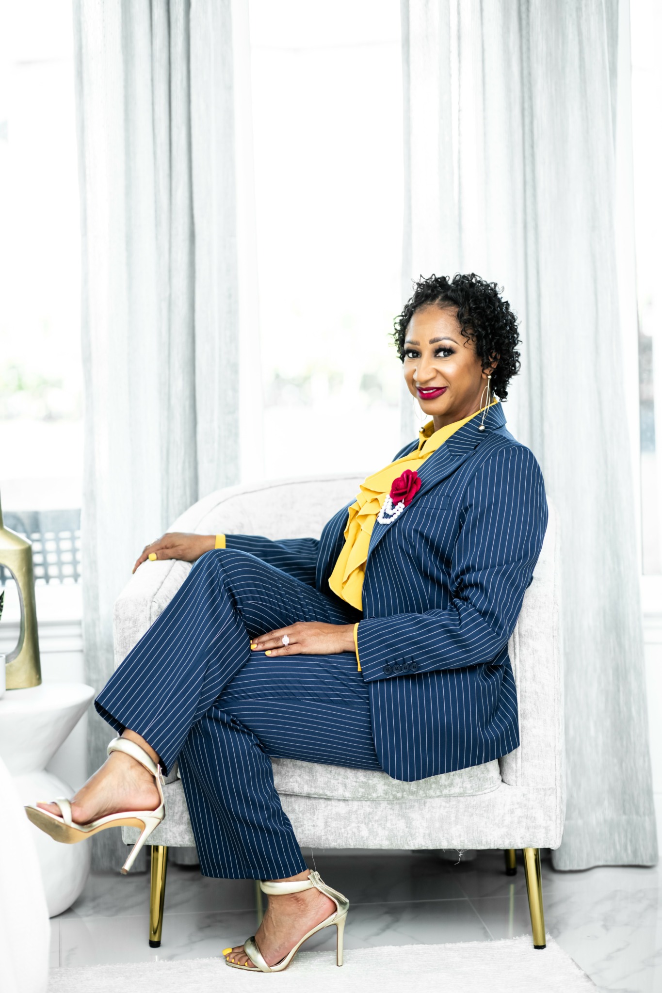 Woman in a blue pinstripe suit sitting on a white sofa, smiling, with a bright window behind her.