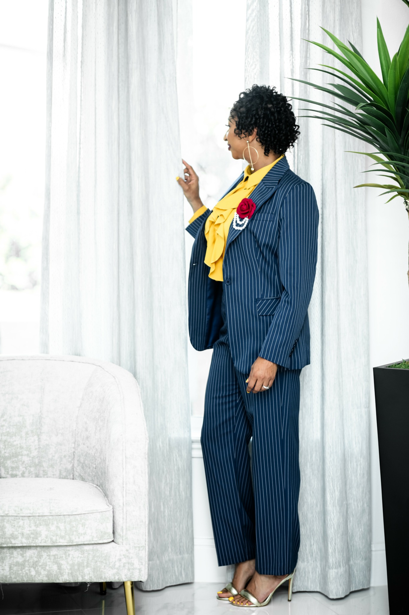 Woman in a pinstripe suit with a yellow scarf and red flower pin, standing near a window with white curtains.