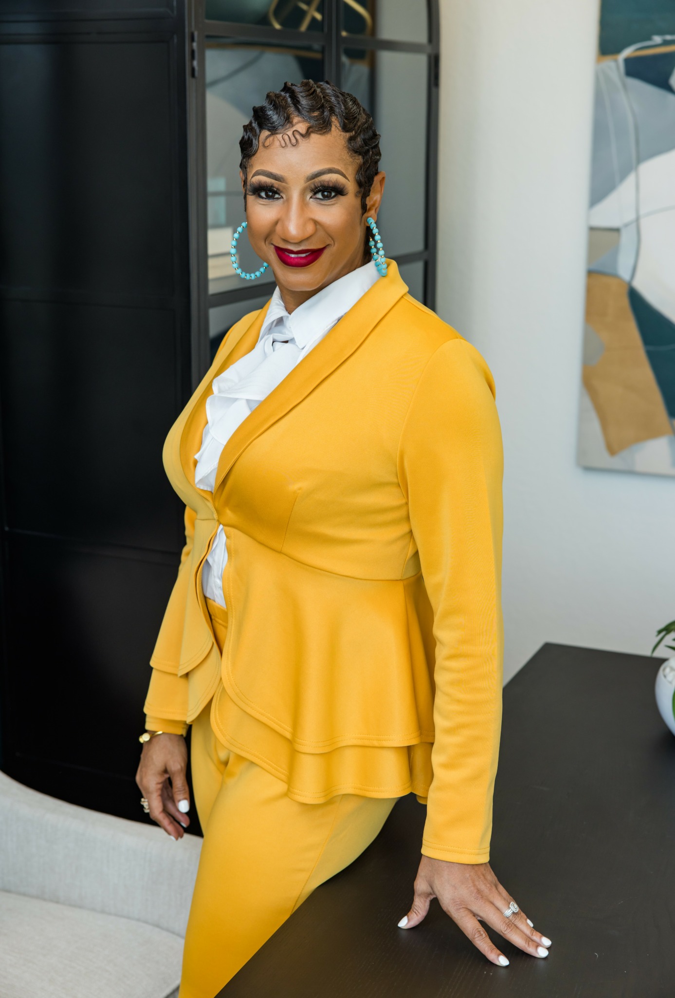 Woman in yellow blazer and pants, smiling, standing indoors near a black cabinet and artwork.
