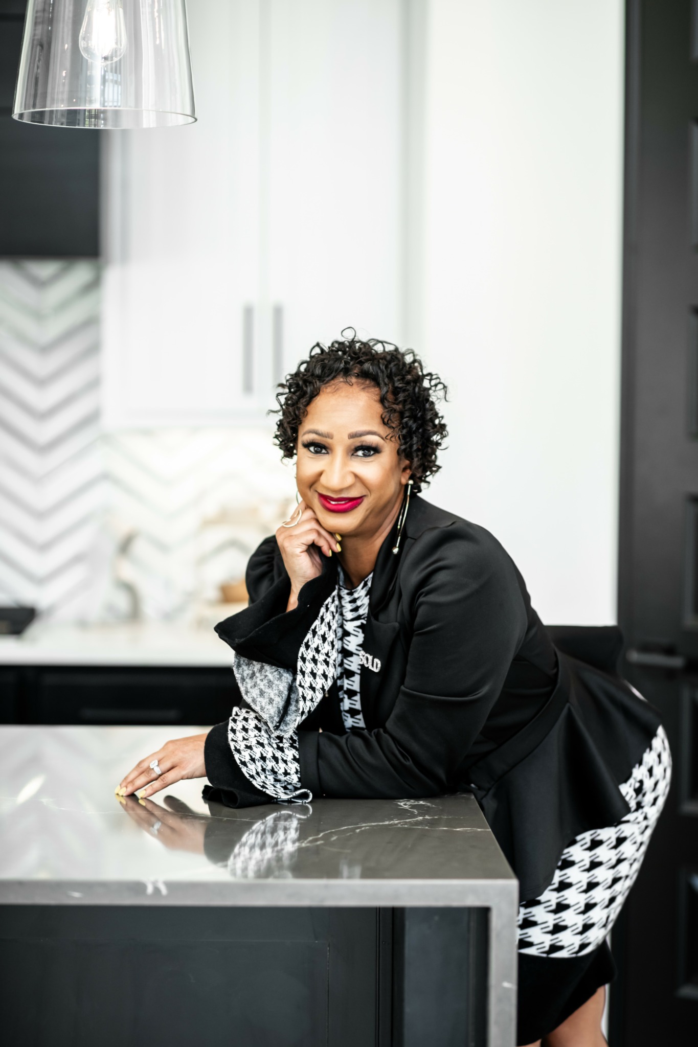 Woman with curly hair and red lipstick leaning on kitchen counter, smiling, wearing black and white patterned clothing.